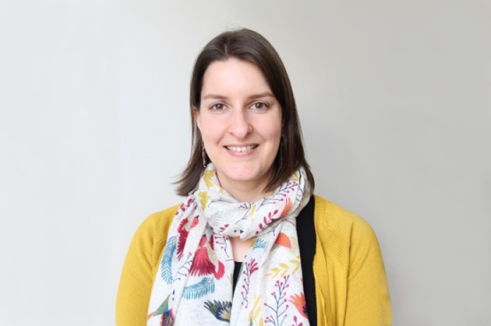 Brown-haired woman in yellow cardigan smiling against plain background