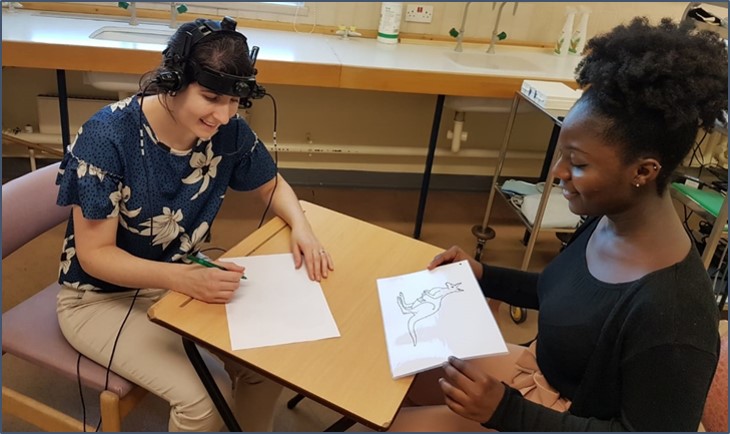 Woman sat down with medical device on head, another sat down holding up a drawing