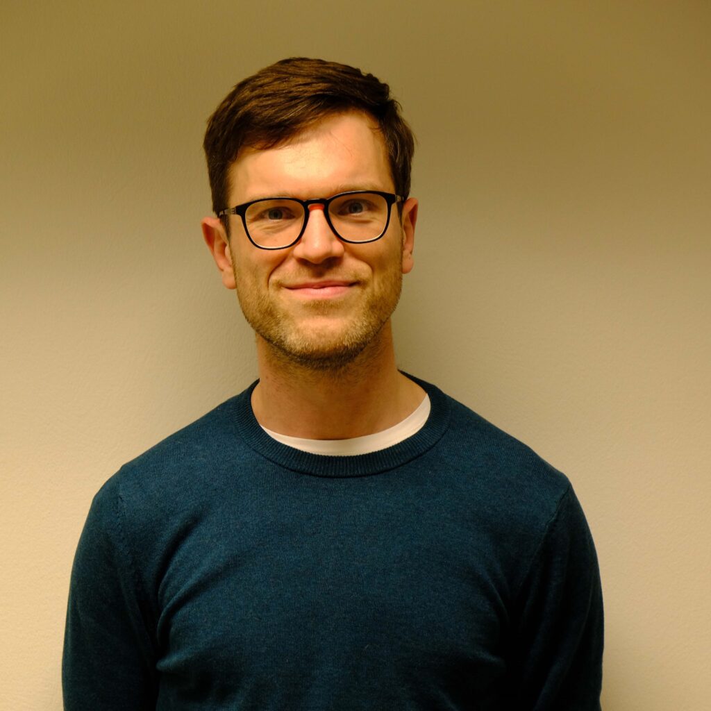 Headshot of Dr Tim Whitfield. Tim is smiling and wearing dark rounded glasses and a blue round-neck jumper.